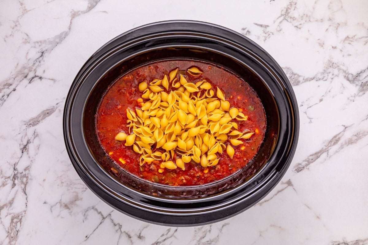 Uncooked shell pasta on top of red sauce in a black slow cooker on marble.