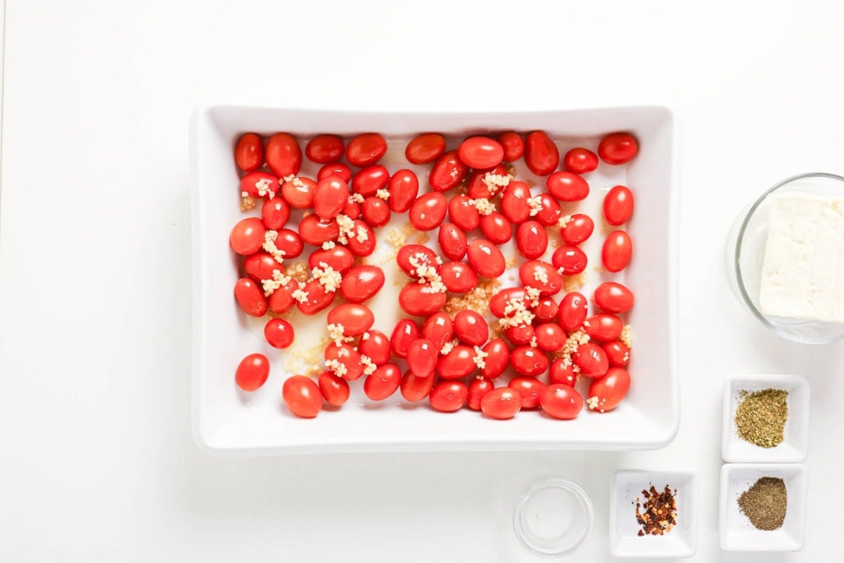 Tomatoes with minced garlic in a dish, spices and feta cheese nearby.