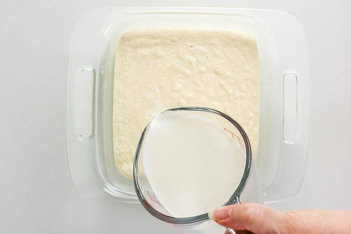 Hand pouring milk from a measuring cup over batter in a square glass dish.