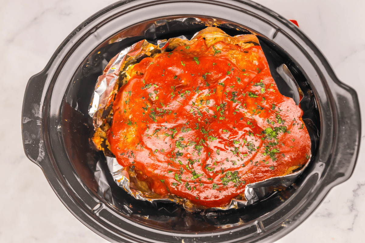 Meatloaf with tomato sauce in a slow cooker, topped with chopped herbs.