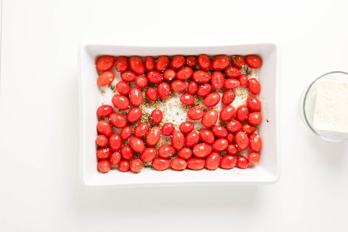 Cherry tomatoes in a white baking dish, block of cheese in a bowl beside it.