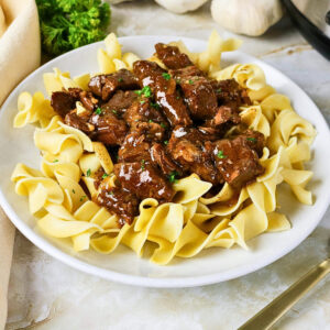 Beef stew served over wide egg noodles on a white plate.