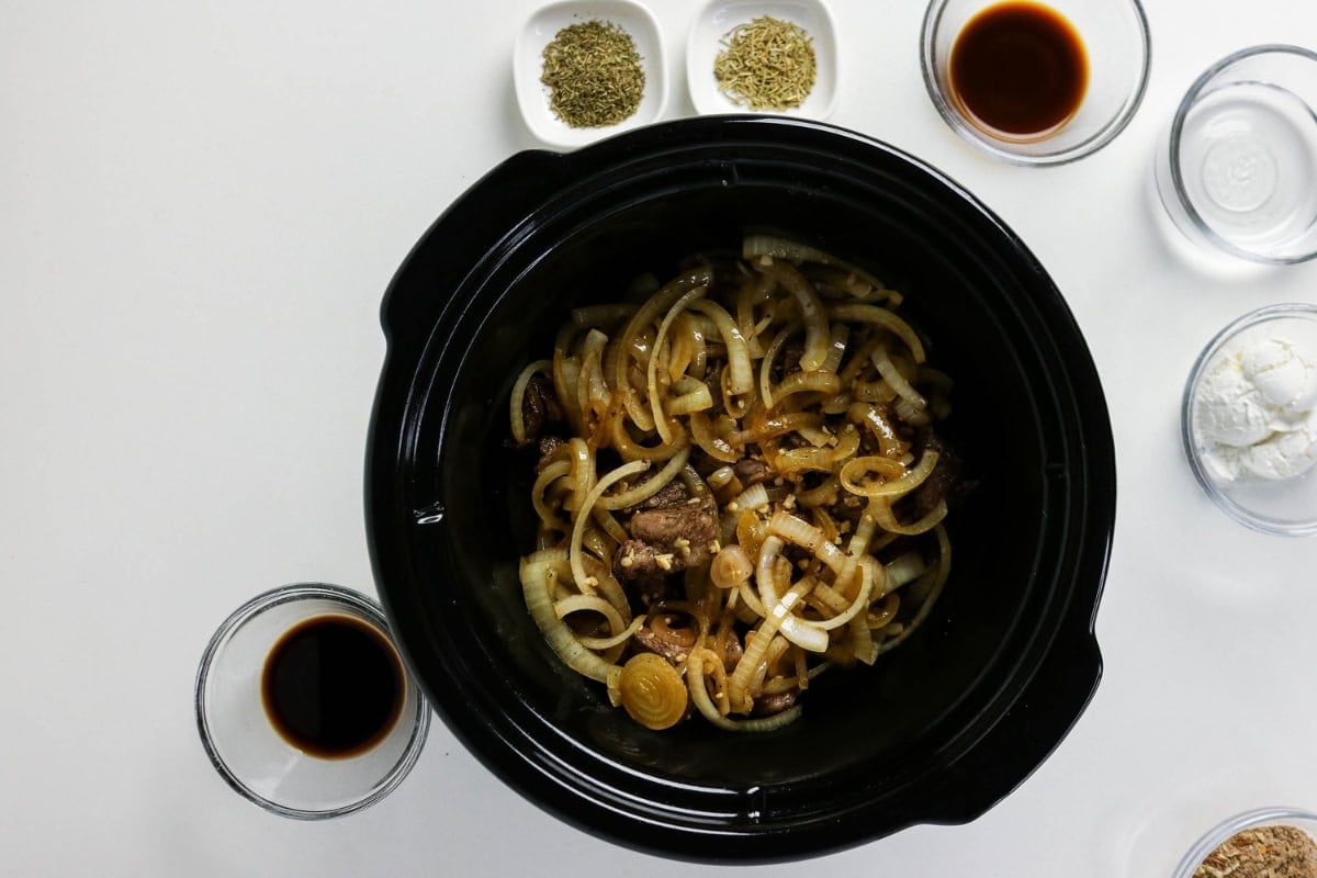 Sliced onions and beef in a slow cooker, surrounded by various seasonings.