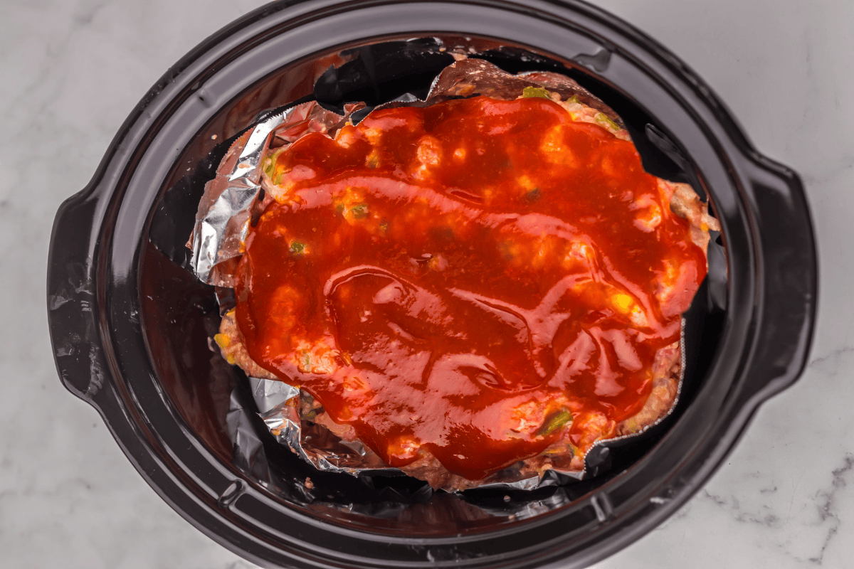 Meatloaf topped with red sauce in a slow cooker, lined with foil.