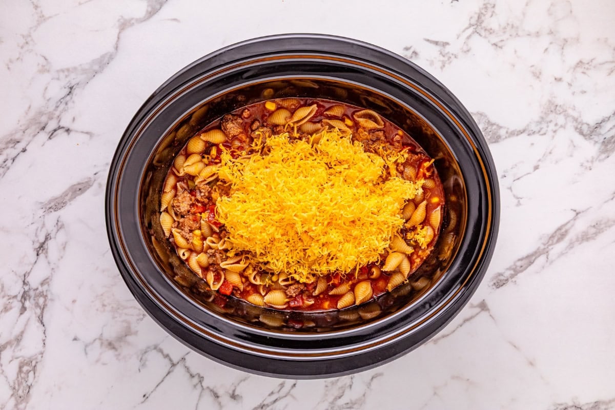 Slow cooker with pasta, meat sauce, and shredded cheese on a marble counter.