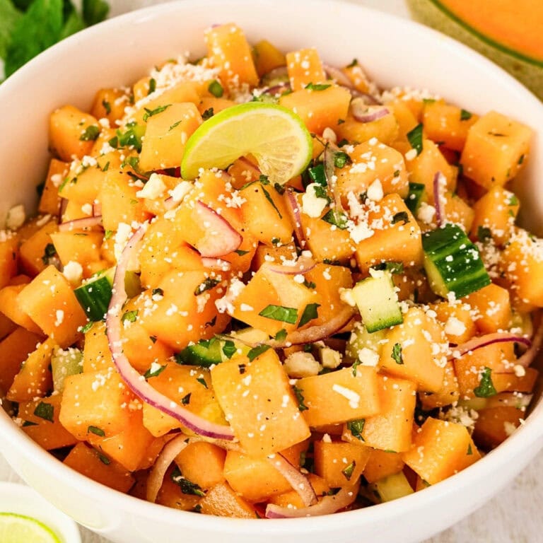 Bowl of cantaloupe salad with cucumber, onion, cheese, herbs, and a lime wedge.