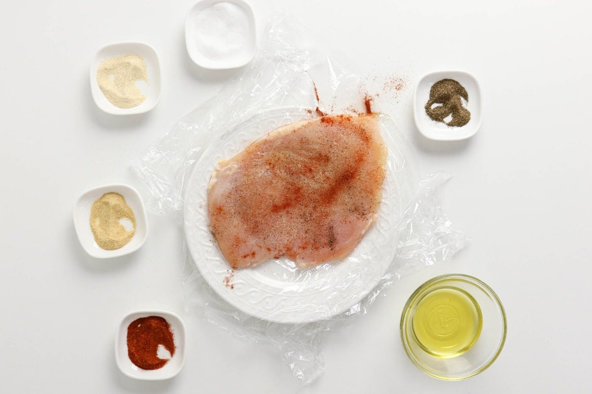 Raw chicken breast on a plate with spices and oil in small bowls around it.