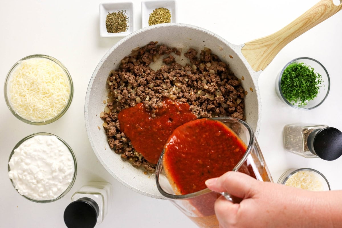 Tomato sauce is poured into browned beef; cheese and herbs are nearby.