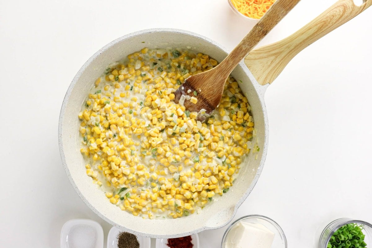 Corn in creamy sauce is stirred in a white pan with a wooden spoon.