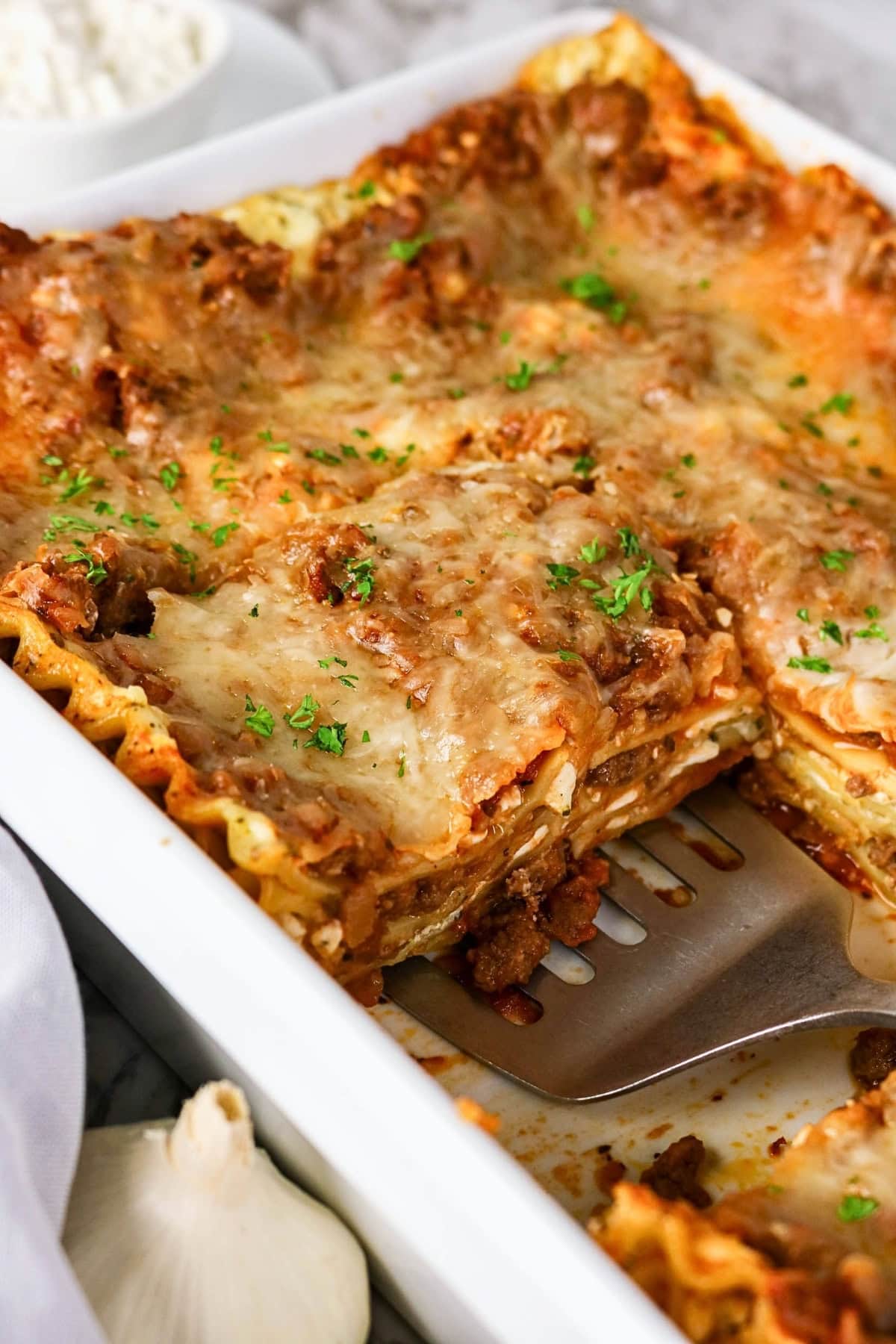 A pan of cheesy baked lasagna with a slice being served.