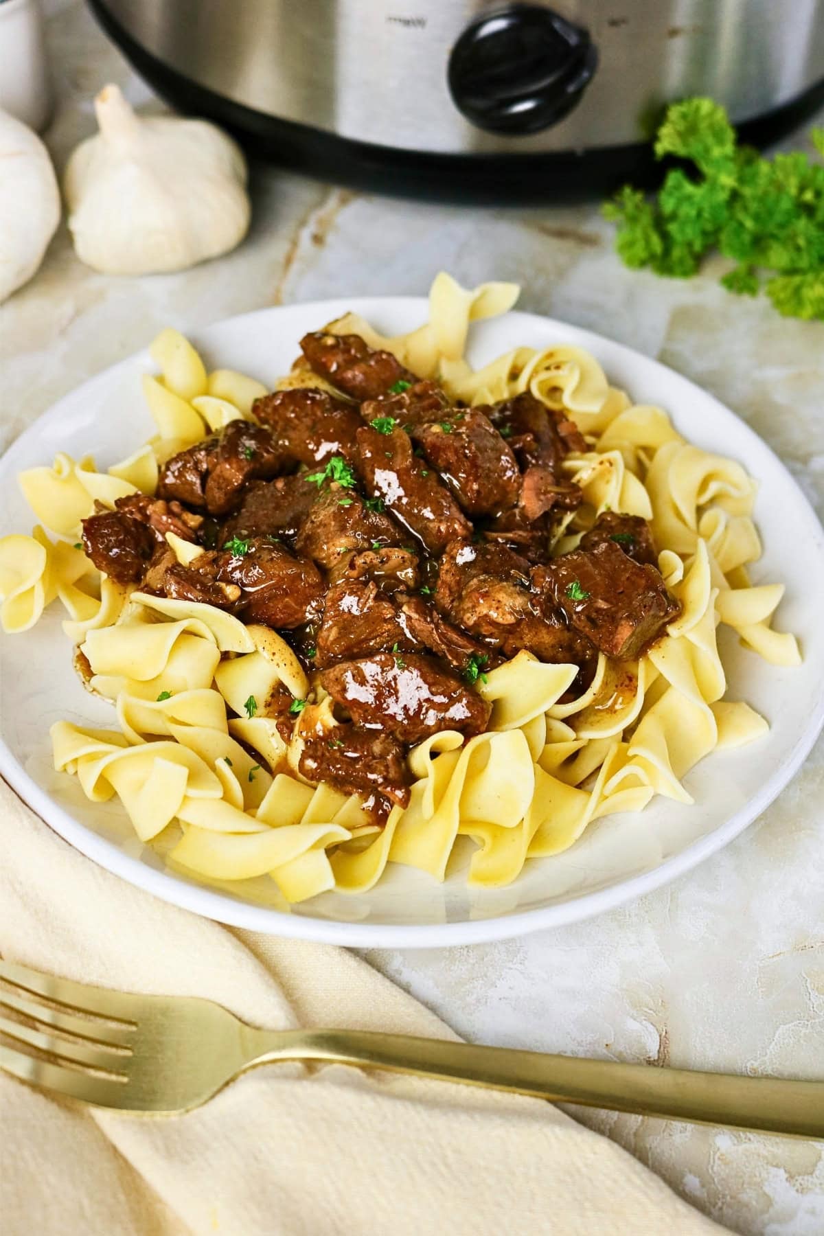 Beef stew over egg noodles on a white plate with a fork and garlic nearby.