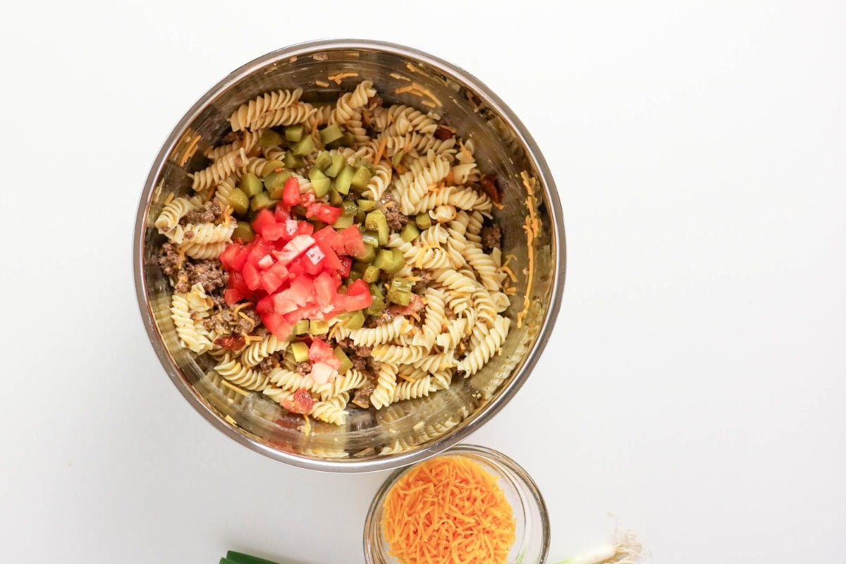 A bowl of rotini pasta salad with diced tomatoes and pickles, cheese nearby.