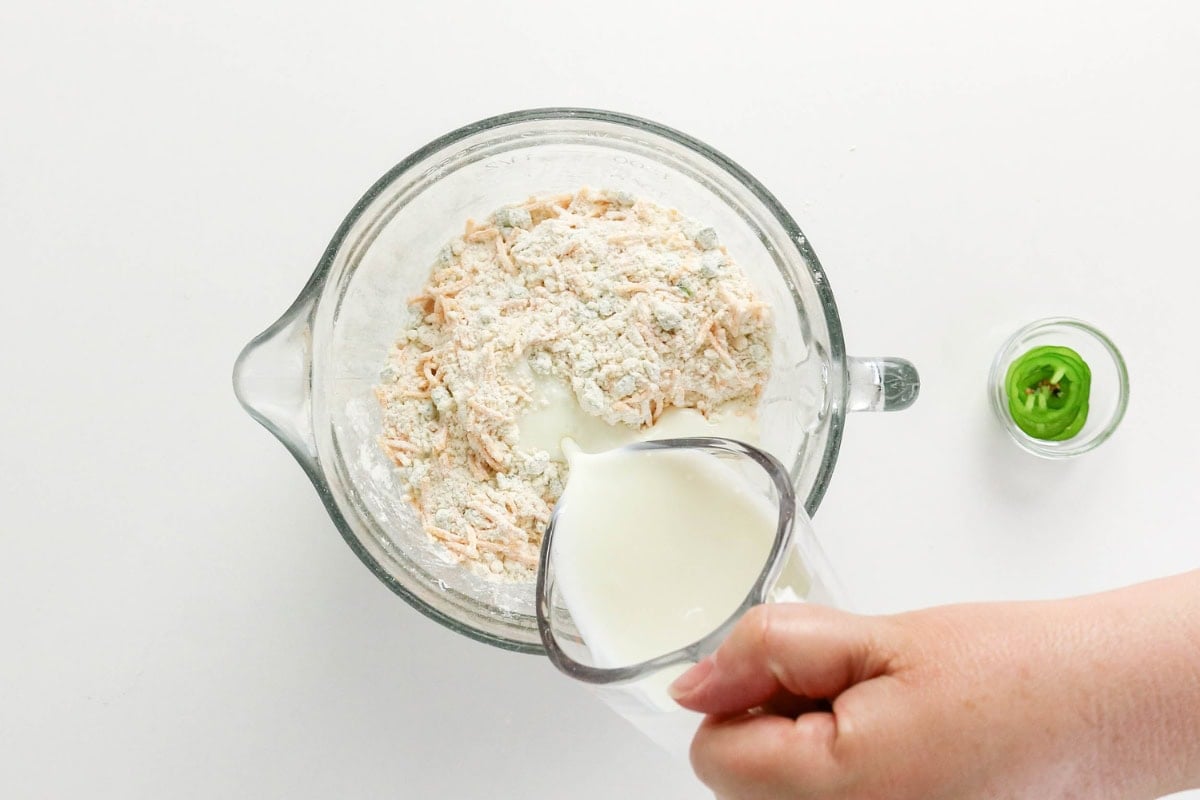 Hand pouring milk into a bowl of flour mixture, jalapeño in small dish nearby.