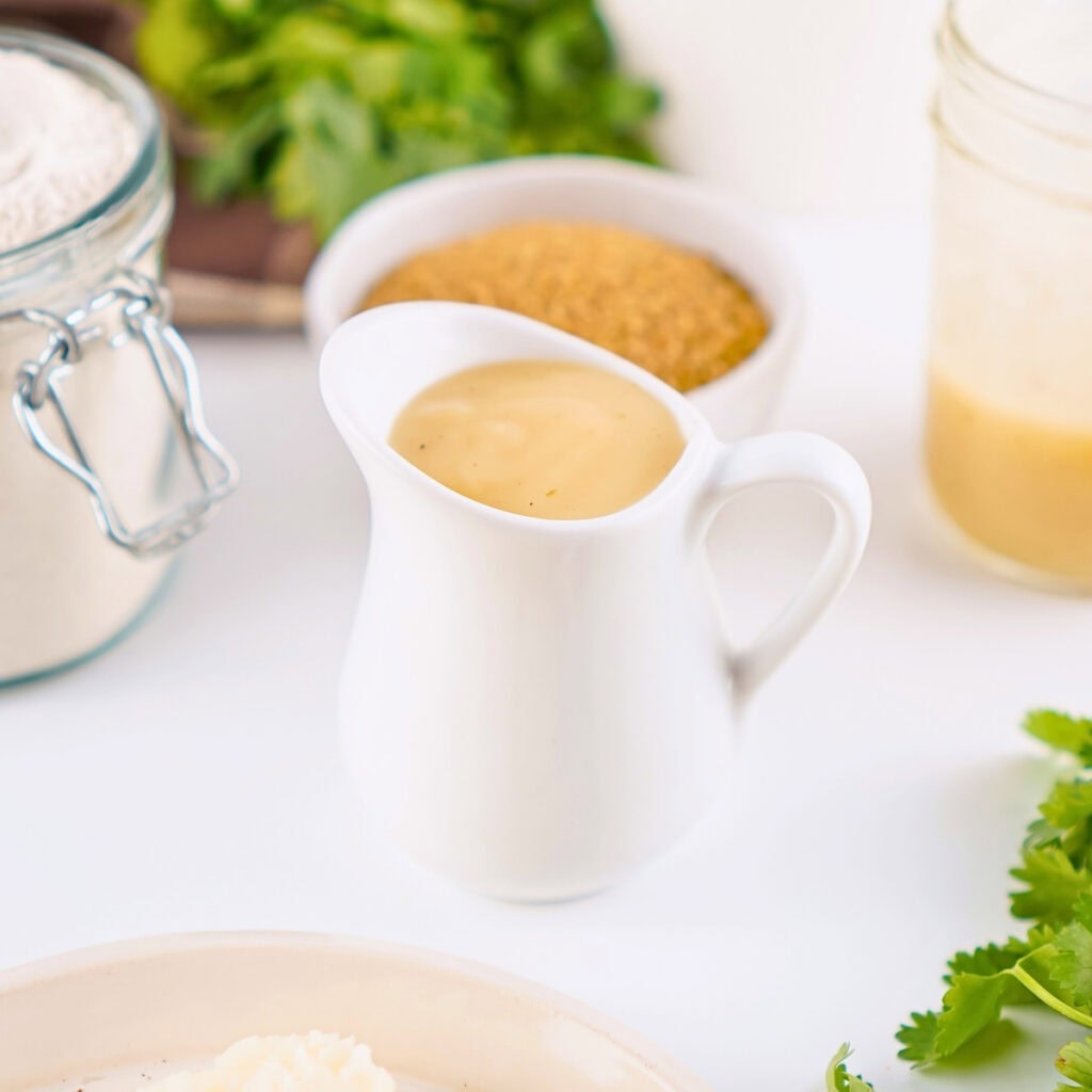 Small white pitcher of gravy on a table with herbs and ingredients nearby.
