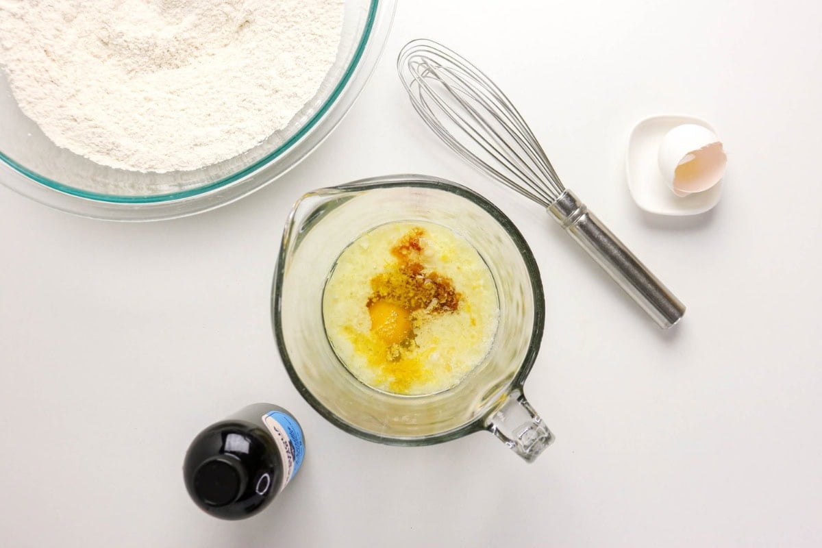 A bowl of flour, whisk, egg mixture, vanilla, and eggshell on a white surface.