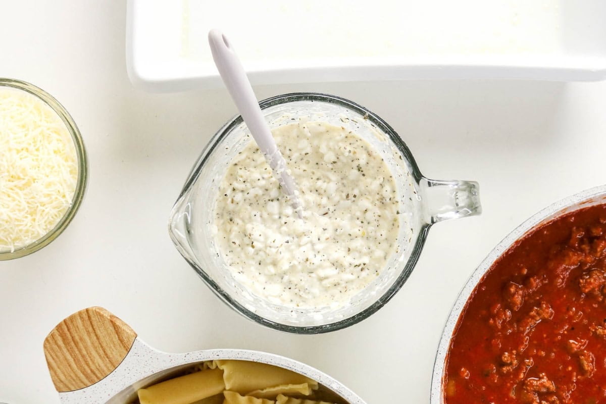 Glass cup with creamy sauce, seasoning, cheese, pasta, and red sauce nearby.