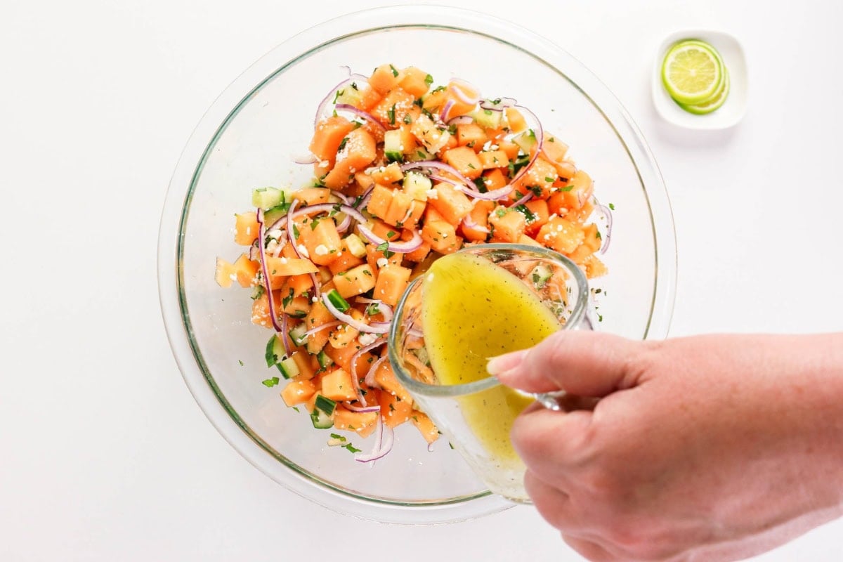 A hand pours dressing onto a fresh tomato and cucumber salad in a glass bowl.