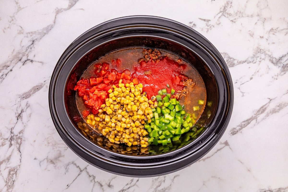 Slow cooker with ground meat, corn, diced tomatoes, and chopped green peppers.