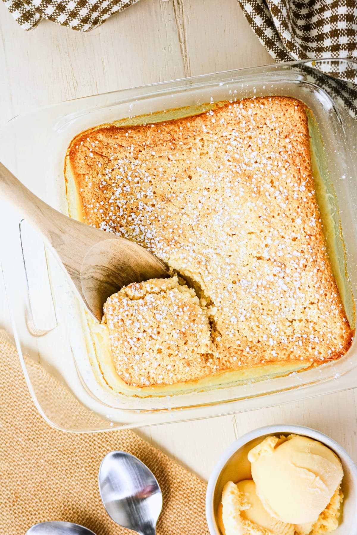 Wooden spoon lifting cake from glass pan, with ice cream and spoons nearby.