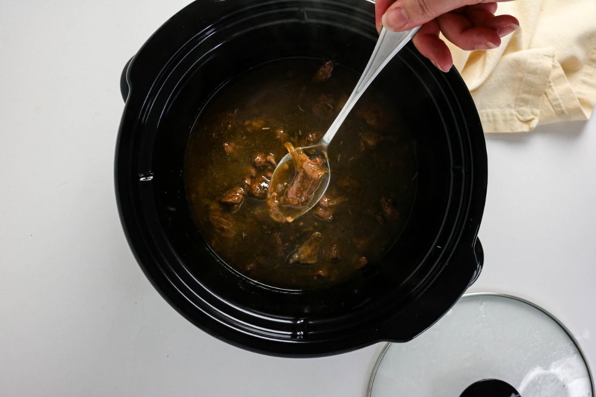 Hand stirring meat stew in a black slow cooker with a metal spoon.