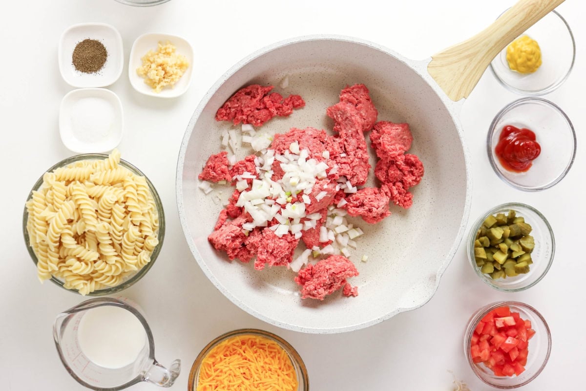 Ground beef and onions in skillet, surrounded by pasta, cheese, and condiments.