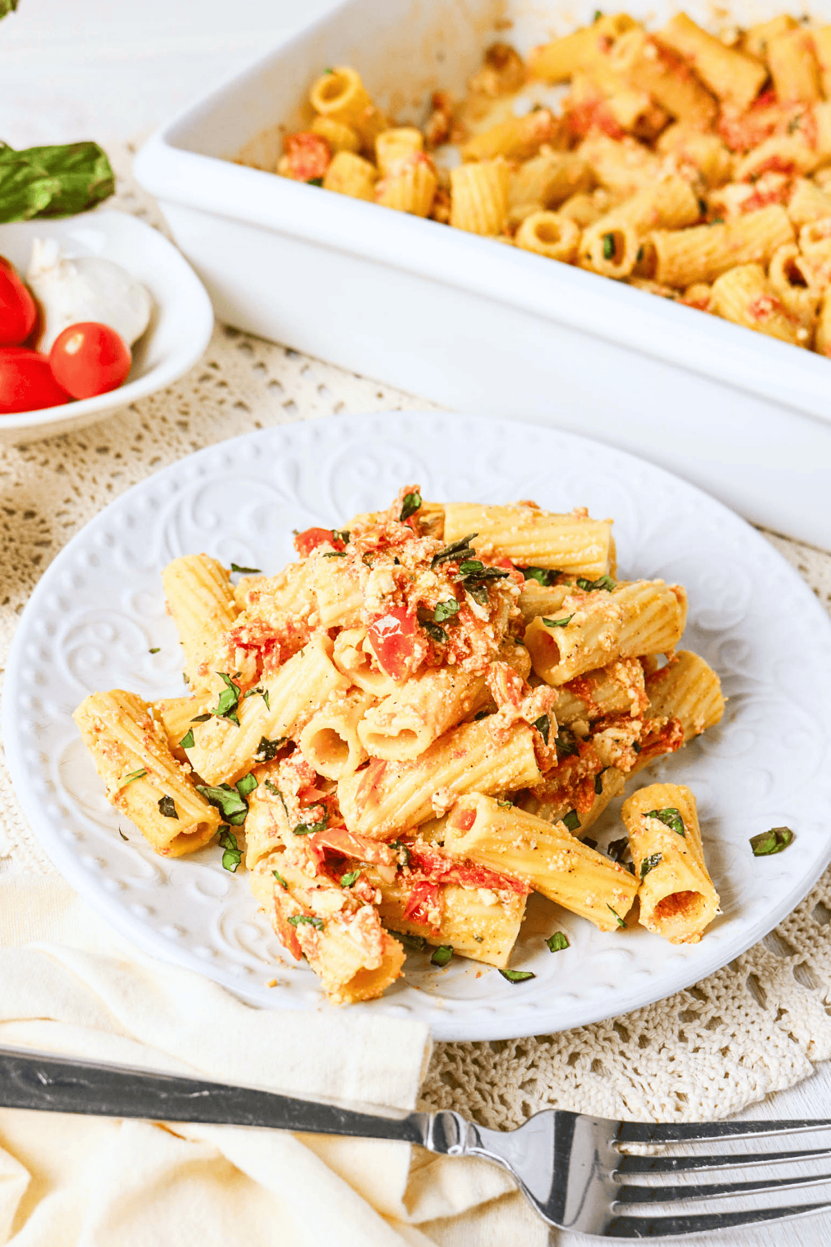 Plate of baked rigatoni pasta with tomato, cheese, and herbs on a white table.