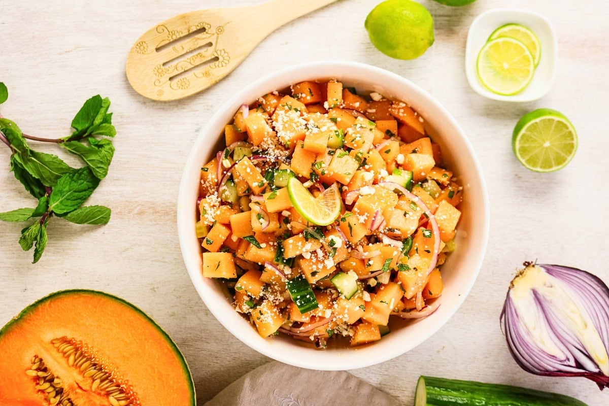 Bowl of cantaloupe salad with lime, cucumber, mint, and cheese on a table.