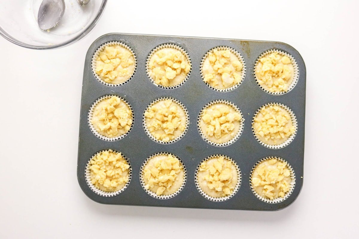 Muffin tin with 12 unbaked crumble-topped muffins on a white background.