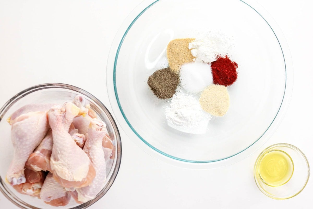 Raw drumsticks, spices in a bowl, and a small bowl of oil on a white surface.