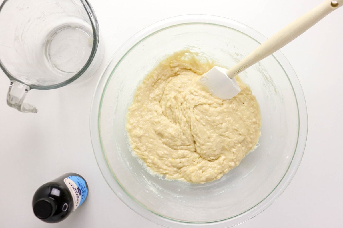 A bowl of batter with a spatula, next to a glass cup and vanilla extract.