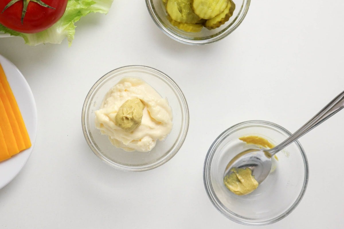 Small bowls of pickles, mayonnaise with mustard, and a spoon on a white table.