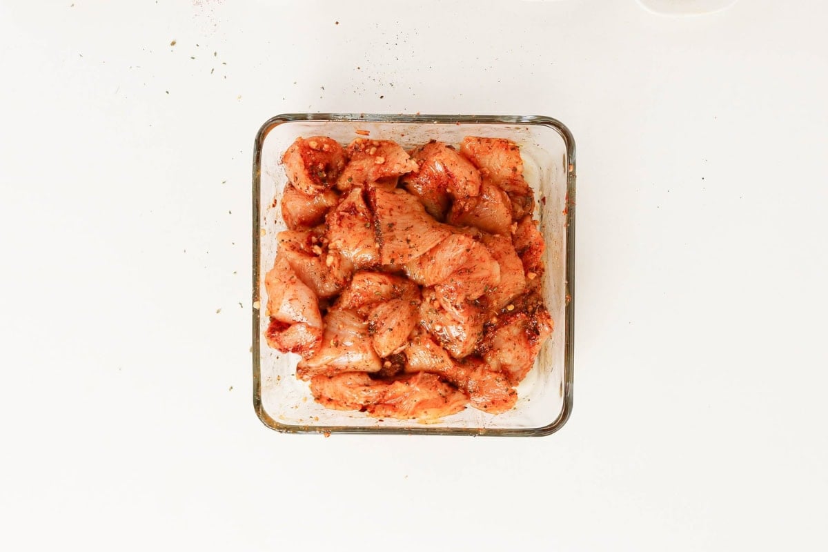 Raw, seasoned chicken pieces in a glass container on a white background.