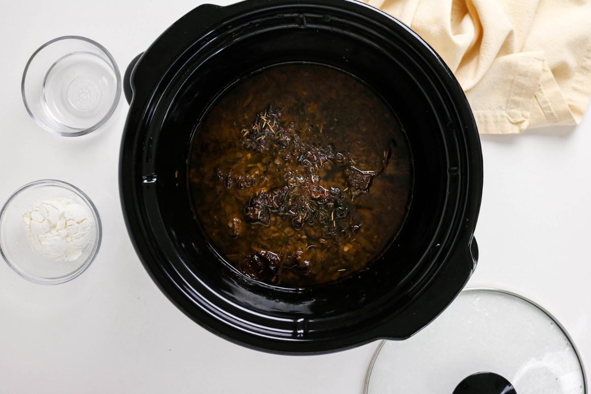 A slow cooker with stew, next to bowls of flour and water, and a towel.