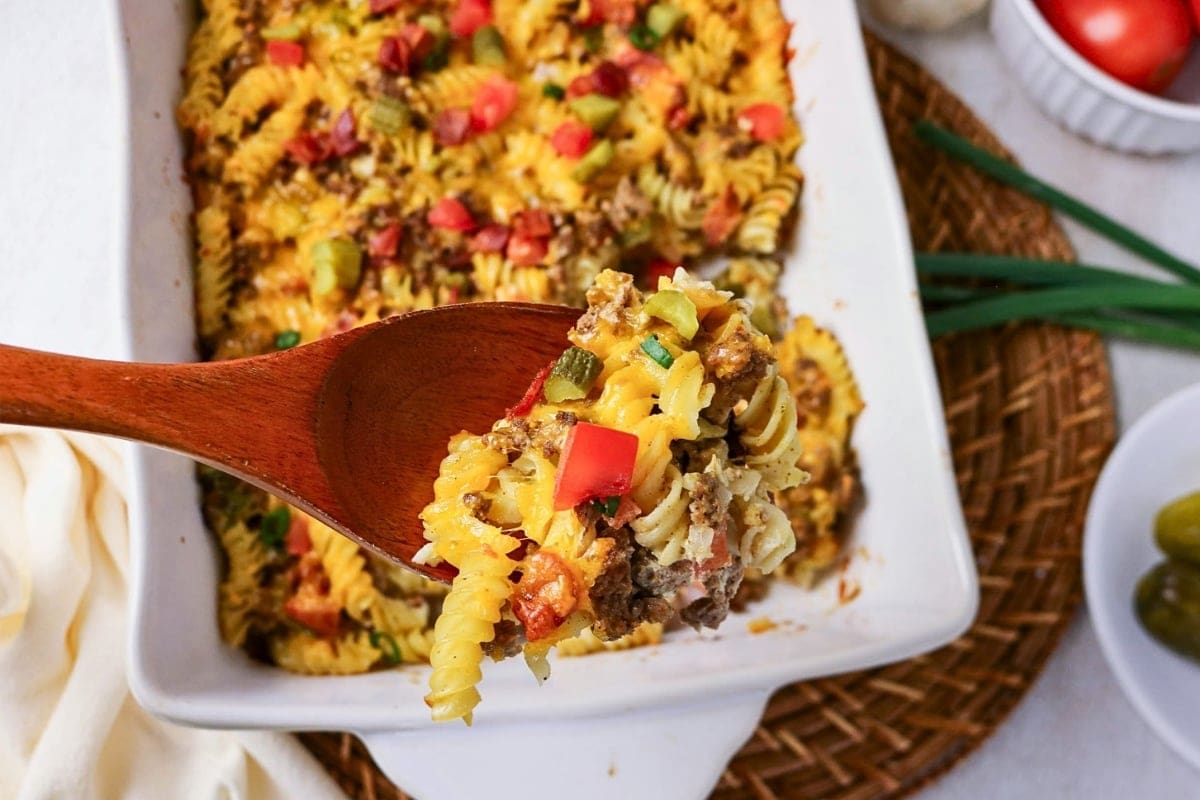 A wooden spoon holds cheesy pasta bake with veggies above a casserole dish.