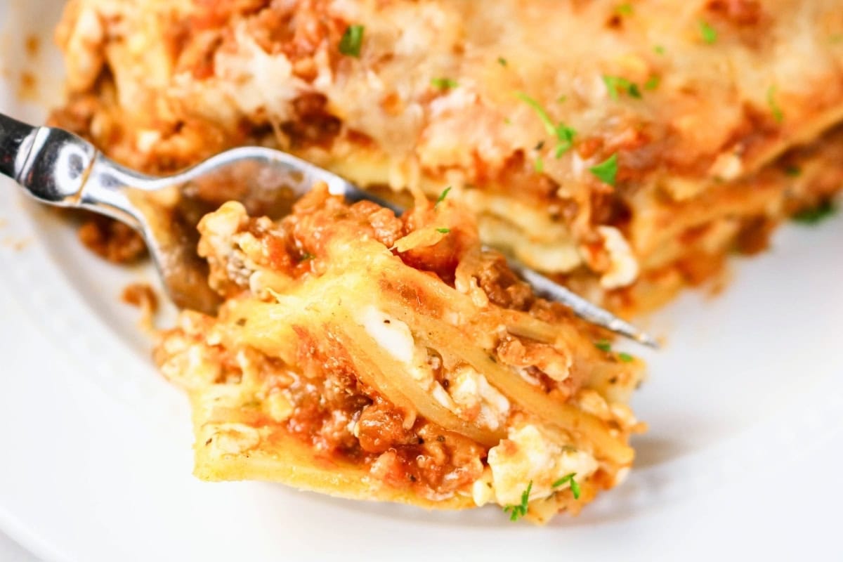 Fork holding a bite of cheesy lasagna over a plate.