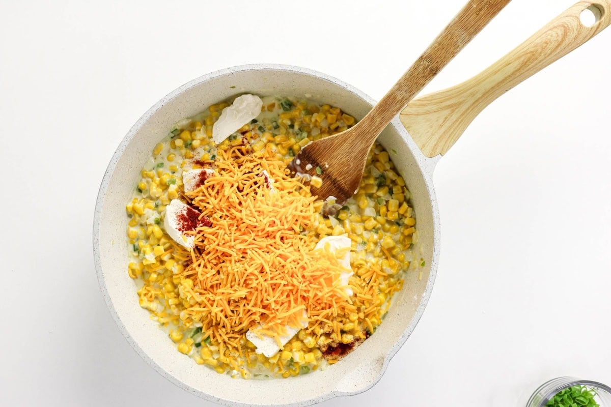 Corn, shredded cheese, and cream in a pan being stirred with a wooden spoon.