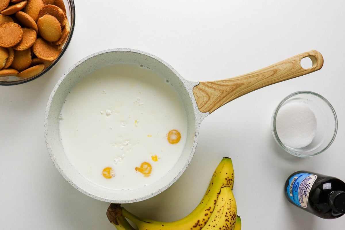 A saucepan with milk, surrounded by bananas, sugar, cookies, and vanilla.