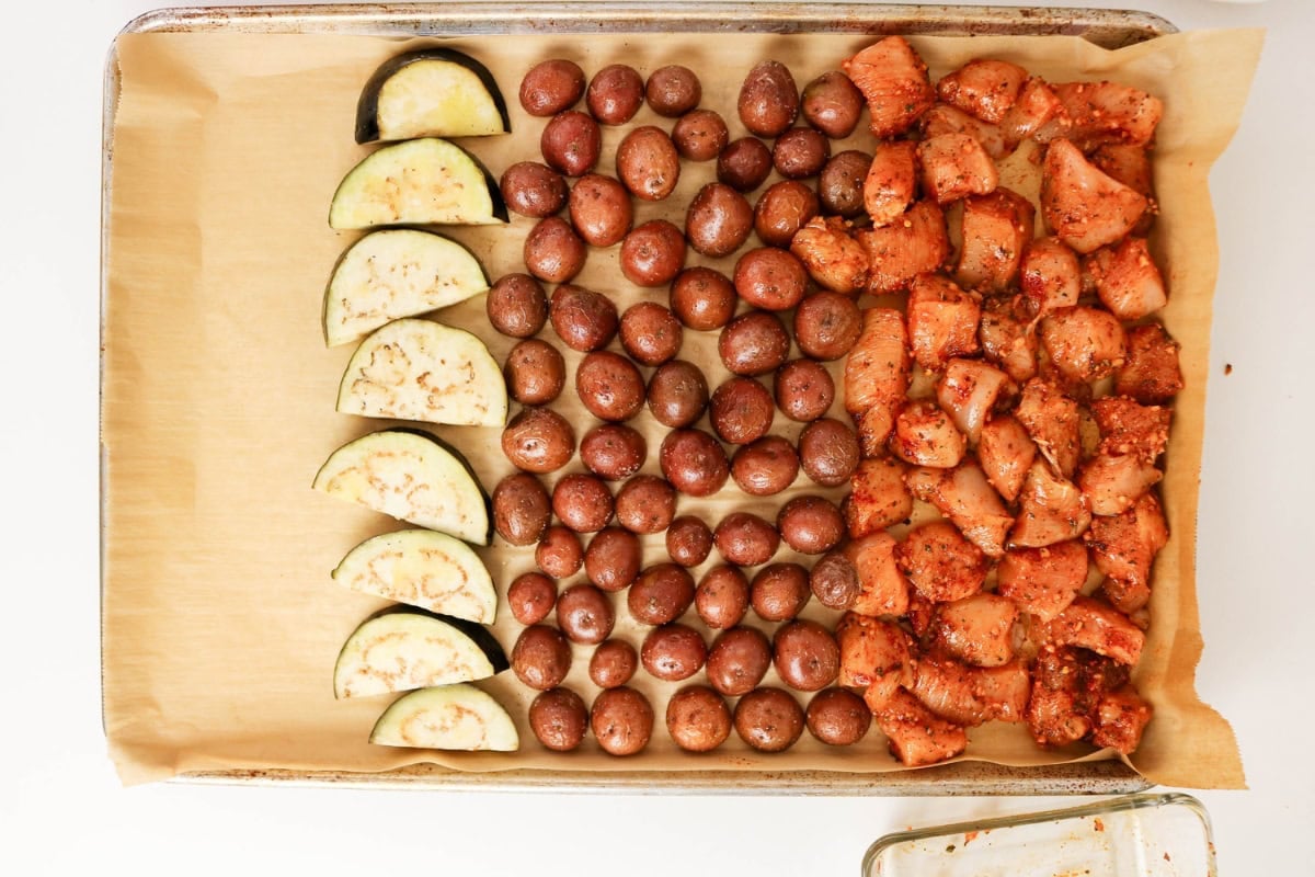 Sliced eggplant, baby potatoes, and seasoned chicken pieces on a baking sheet.