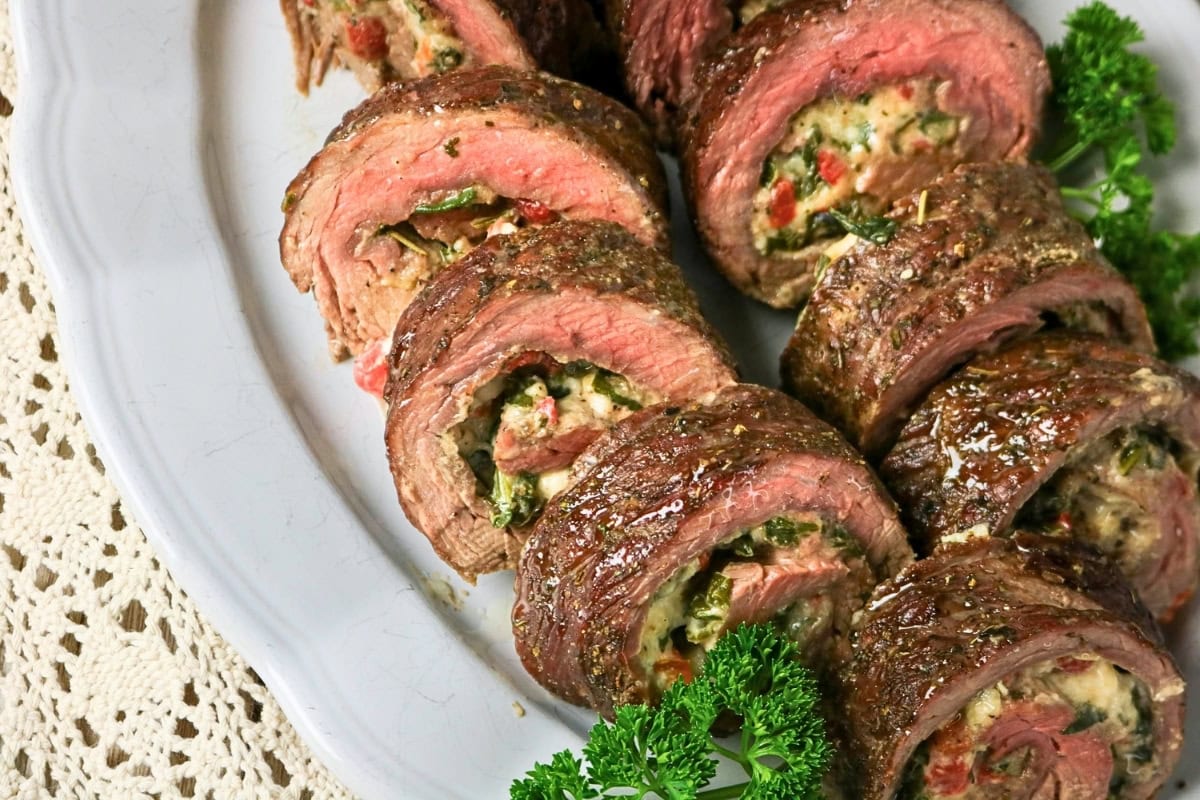 Sliced stuffed steak rolls with herbs and vegetables on a white platter.
