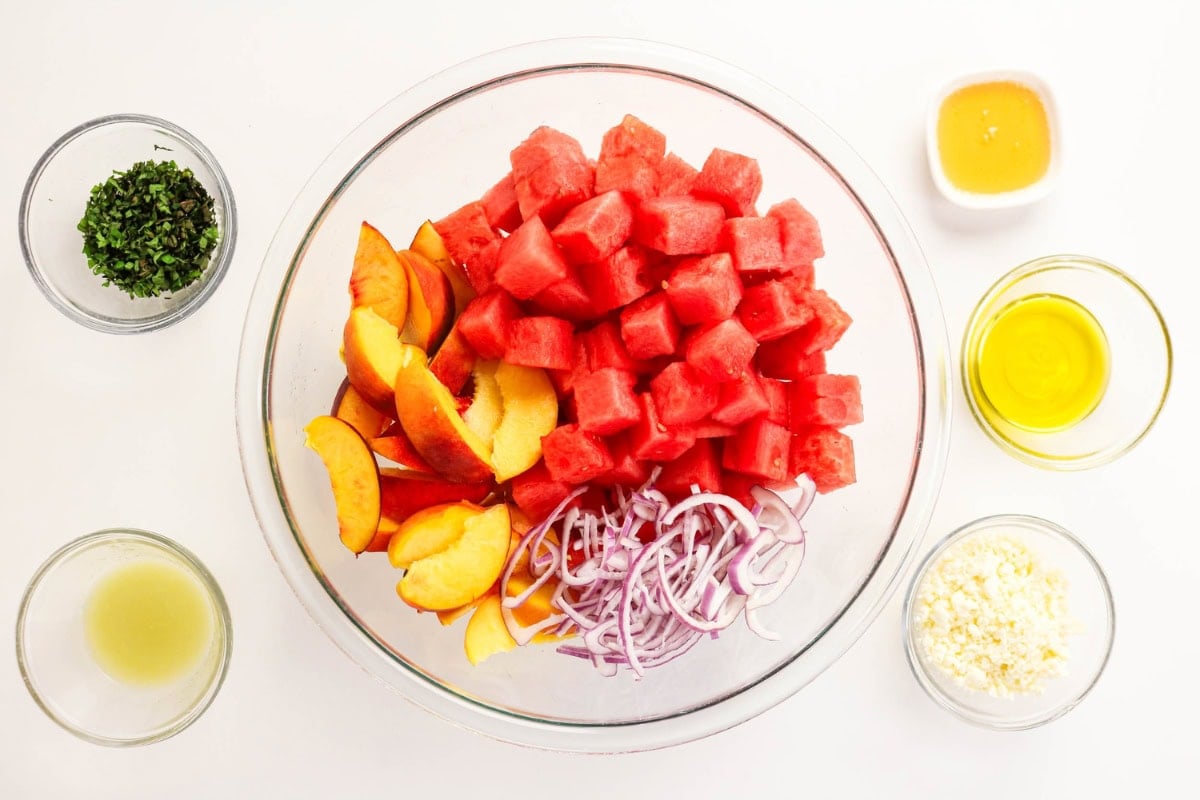 Glass bowl with watermelon, peach, red onion; small bowls with toppings nearby.