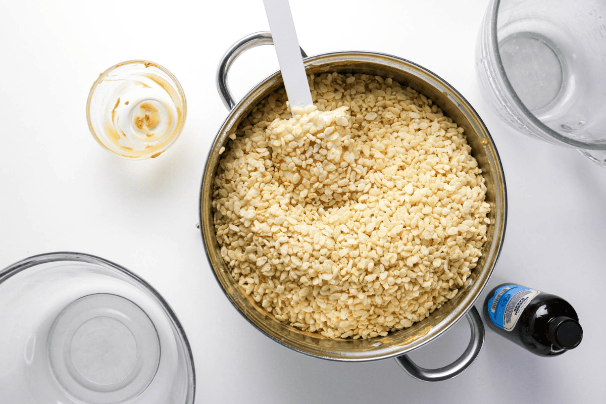 Pot of rice cereal with a spatula, vanilla, and empty bowls on a white table.