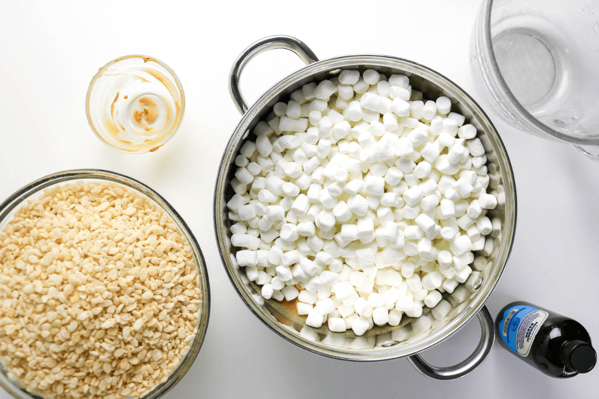 Mini marshmallows, rice cereal, vanilla, and a glass bowl on a white surface.