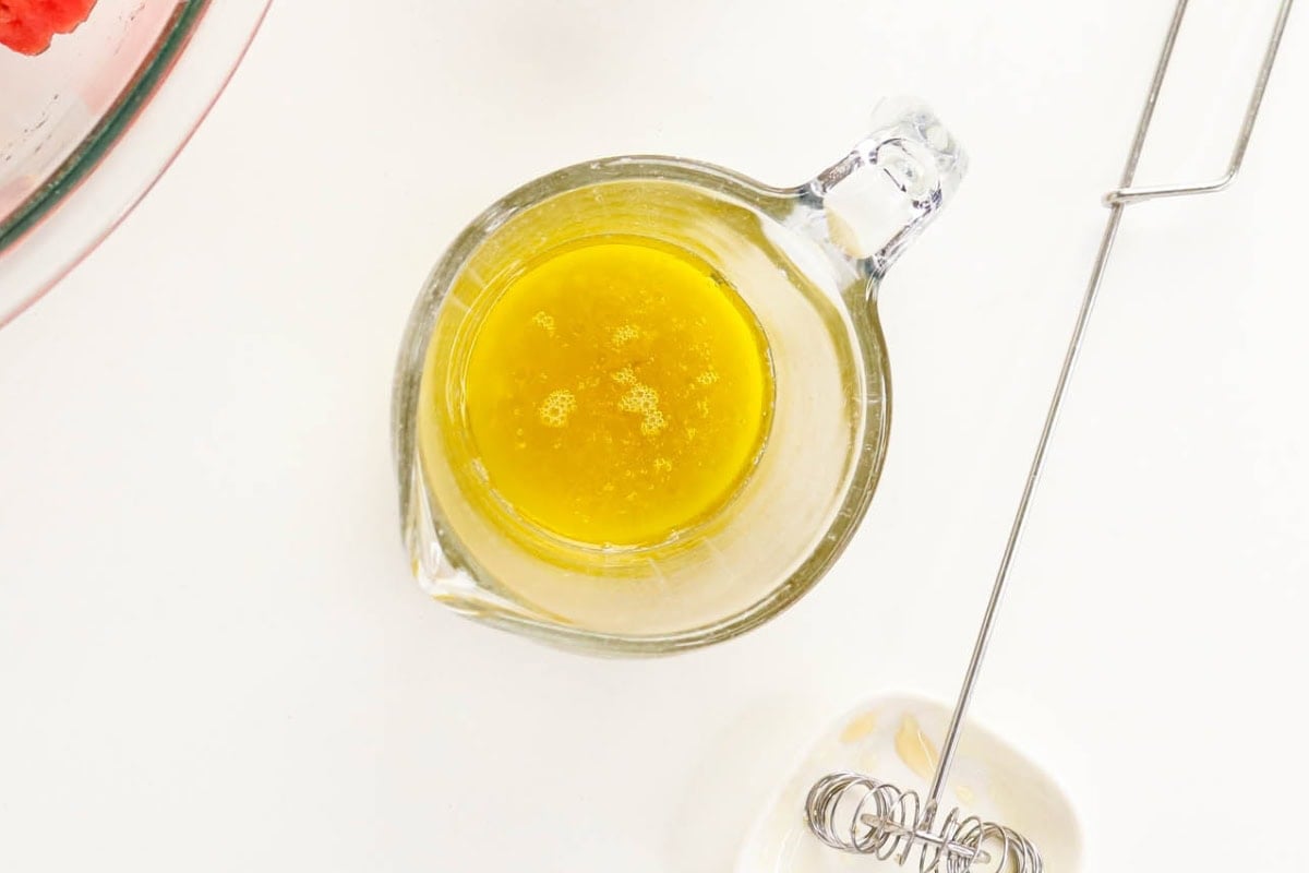 Glass measuring cup with yellow liquid beside a whisk on a white surface.