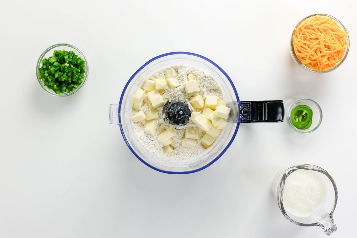 Food processor with flour and butter, cheese, peppers, and milk in bowls nearby.