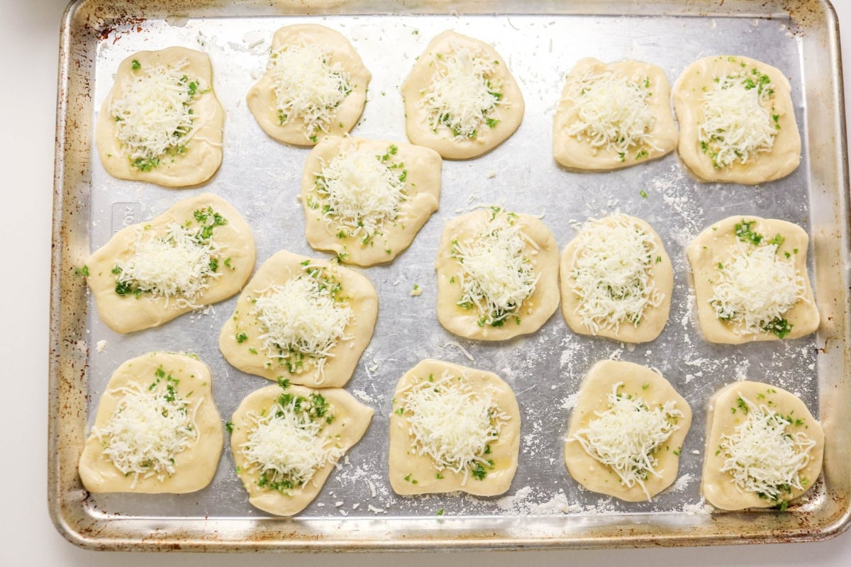 Unbaked dough rounds topped with cheese and herbs on a metal baking sheet.