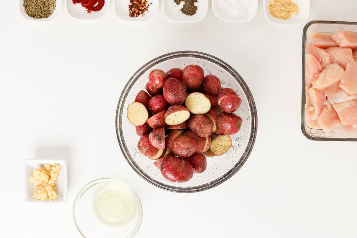 A bowl of halved red potatoes surrounded by spices, oil, and chicken pieces.