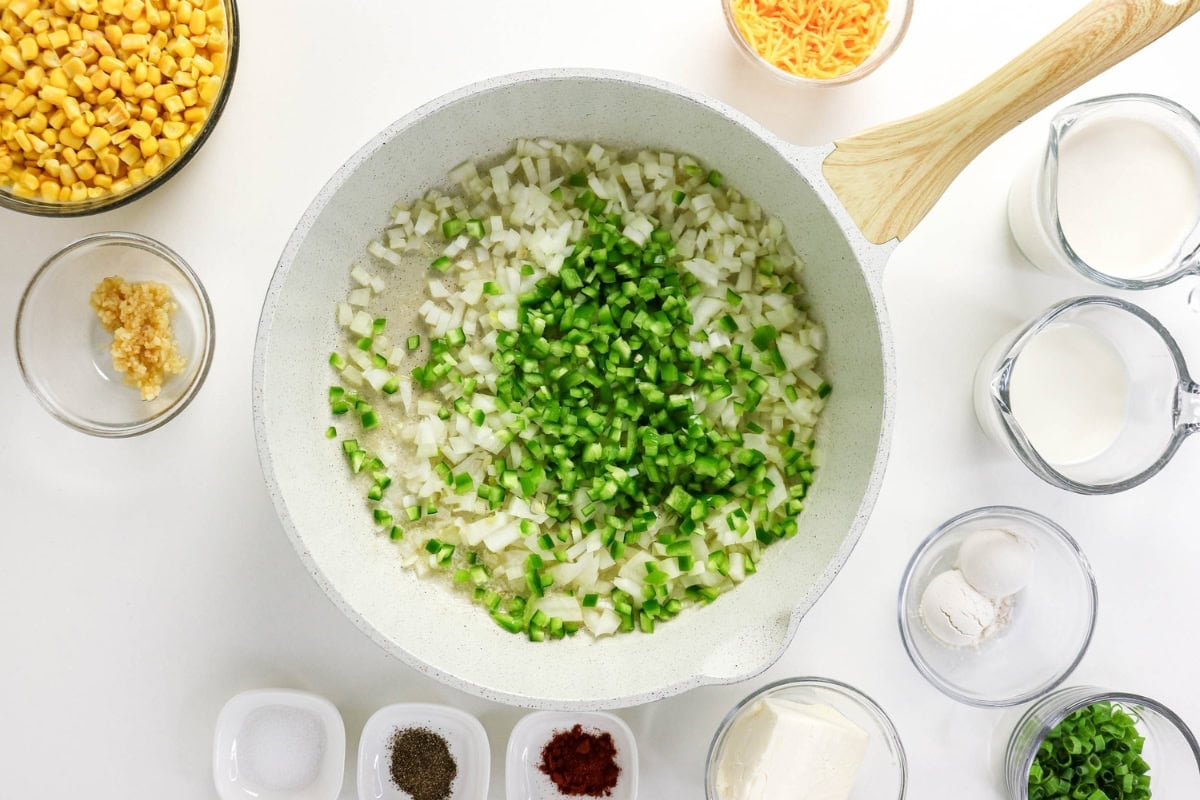 Chopped onions and peppers cooking in a white pan, surrounded by ingredients.