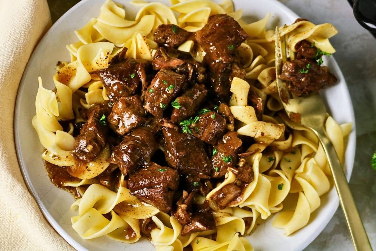 Beef stew served over egg noodles with a fork on a white plate.