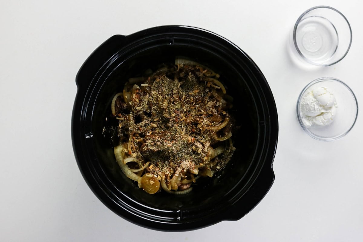 Sliced onions and herbs in a black slow cooker with two bowls on the side.