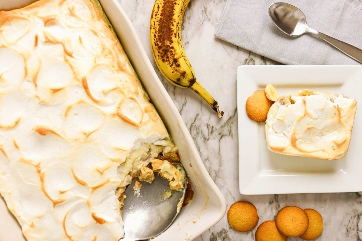 Banana pudding in a dish with a serving on a plate, banana, and cookies nearby.